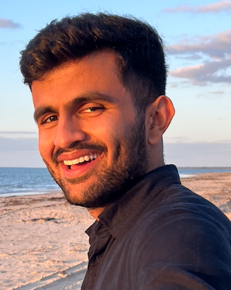 Kushagra Rathore has short dark brown hair and a beard and is wearing a black shirt.  Kushagra is standing outside at the beach.